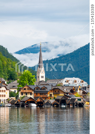 Hallstatt Village and Hallstatter See lake in Austria. Scenery with famous old church near the lake. Clouds and mist over the mountains in background. Famous tourist destination. Hallstatt Village and Hallstatter See lake in Austria. Scenery with famous old church near the lake. Clouds and mist over the mountains in background. Famous tourist destination. 135410389
