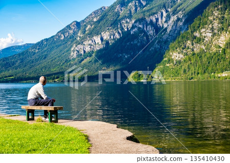 Unidentifiable old men is sitting on bench near the lake in Austrian alps. Concept of relaxation and meditation near nature. Hallstatt city, Austria. Unidentifiable old men is sitting on bench near the lake in Austrian alps. Concept of relaxation and meditation near nature. Hallstatt city, Austria. 135410430