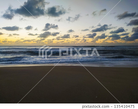 A dramatic morning seascape where rolling blue waves meet dark sands under a cloudy, golden horizon. This tranquil scene evokes a sense of renewal, perfect for wellness or travel content. 135410475