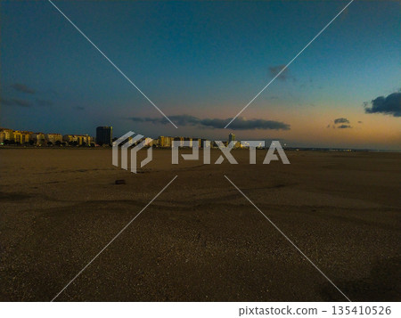 Serene coastal city skyline at dusk across a vast sandy beach. The vibrant sunset sky blends urban life with nature, ideal for travel, real estate, and peaceful background concepts. 135410526