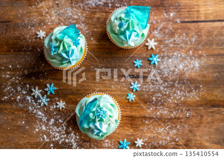 Homemade cupcake of blue color placed on wooden desk. Winter and christmas theme, snowflakes on cream with blue ice. Fresh and home baked sweet. Homemade cupcake of blue color placed on wooden desk. Winter and christmas theme, snowflakes on cream with blue ice. Fresh and home baked sweet. 135410554