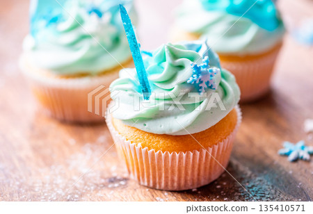 Homemade cupcake of blue color placed on wooden desk. Winter and christmas theme, snowflakes on cream with blue ice. Fresh and home baked sweet. Homemade cupcake of blue color placed on wooden desk. Winter and christmas theme, snowflakes on cream with blue ice. Fresh and home baked sweet. 135410571