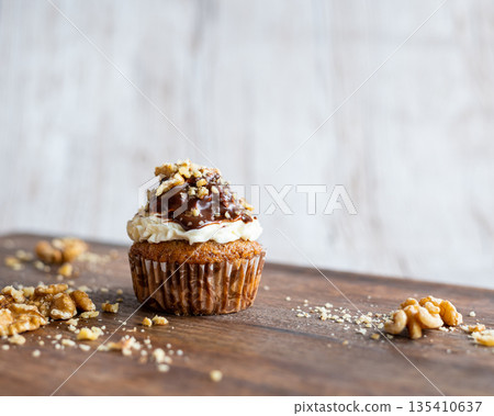Tasty chocolate muffin with creme mascarpone and chocolate on top, covered with nuts. Placed on wooden desk. Sweet fresh and homemade cupcake. 135410637