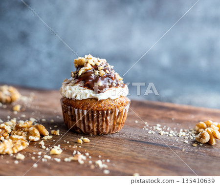 Tasty chocolate muffin with creme mascarpone and chocolate on top, covered with nuts. Placed on wooden desk. Sweet fresh and homemade cupcake. 135410639