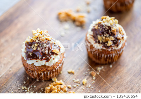 Tasty chocolate muffin with creme mascarpone and chocolate on top, covered with nuts. Placed on wooden desk. Sweet fresh and homemade cupcake. Tasty chocolate muffin with creme mascarpone and chocolate on top, covered with nuts. Placed on wooden desk. Sweet fresh and homemade cupcake. 135410654