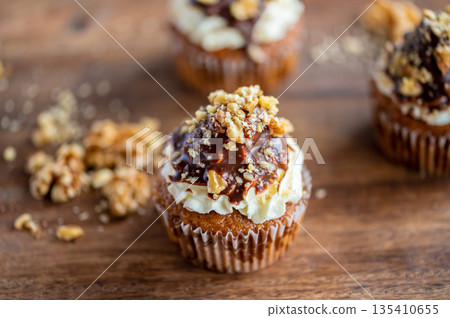 Tasty chocolate muffin with creme mascarpone and chocolate on top, covered with nuts. Placed on wooden desk. Sweet fresh and homemade cupcake. 135410655