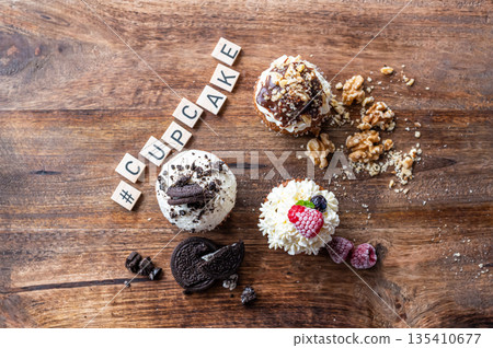 Three tasty cupcakes placed on wooden desk. White creme, chocolate and raspberry, covered with nut. The word cupcake made from wooden blocks. 135410677