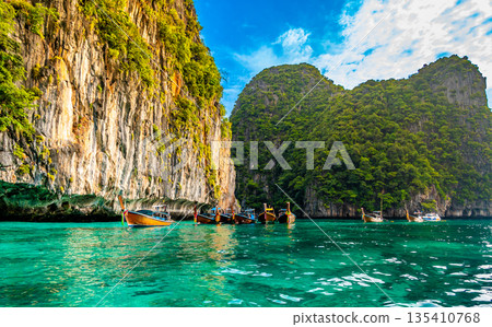 View of long tail boats on turquoise water at ocean near Ko Phi Phi islands, Thailand. Concept of exotic vacation in tropical paradise. Limestone rock in background. View of long tail boats on turquoise water at ocean near Ko Phi Phi islands, Thailand. Concept of exotic vacation in tropical paradise. Limestone rock in background. 135410768