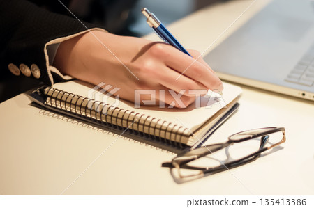 Close up on woman's hands writing in notepad and working on laptop. Freelancer workspace at home or workplace desk at office. Work from home concept. 135413386