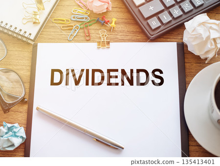 On a wooden table there is an office sheet of paper with the text DIVIDENDS. Business workspace with calculator, glasses, pen, crumpled paper and cup of coffee. 135413401