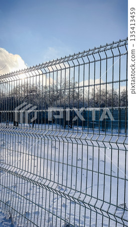 Metal wire fence covered in thick transparent ice glaze against a snowy park and sunny winter sky 135413459