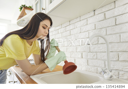 Portrait of a young upset brunette woman cleaning pipe with a cup plunger in the kitchen at home. 135414894