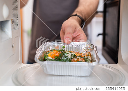 Man placing foil tray with fried eggs into microwave, highlighting unsafe appliance use. Man placing foil tray with fried eggs into microwave, highlighting unsafe appliance use. 135414955
