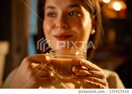Close up of woman holding glass cup of latte in warm cafe lighting. Close up of woman holding glass cup of latte in warm cafe lighting. 135415418