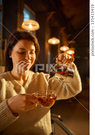 Woman pouring milk into glass cup of coffee in warm cafe interior Woman pouring milk into glass cup of coffee in warm cafe interior 135415419