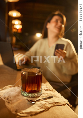 Coffee glass on wooden table with woman smiling and holding smartphone in cafe. Coffee glass on wooden table with woman smiling and holding smartphone in cafe. 135415421