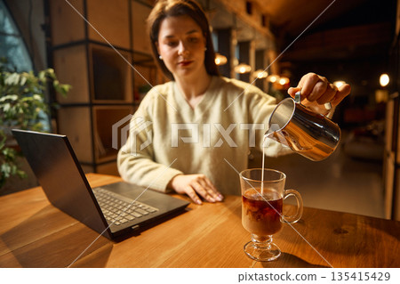 Woman pouring milk into glass coffee cup beside laptop on table. Woman pouring milk into glass coffee cup beside laptop on table. 135415429