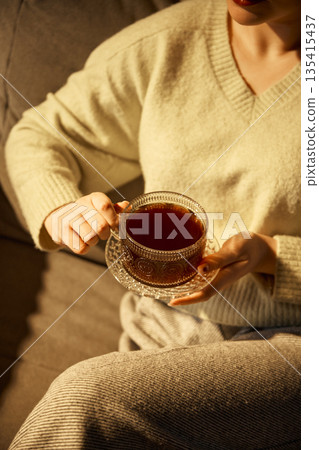 Woman holding glass coffee cup while sitting on sofa indoors. Woman holding glass coffee cup while sitting on sofa indoors. 135415437