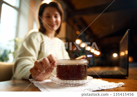 Coffee cup on saucer with woman hand reaching across wooden table. Coffee cup on saucer with woman hand reaching across wooden table. 135415456