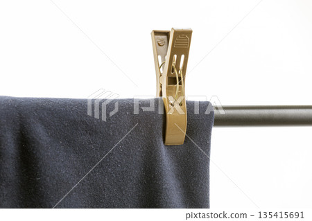 A close-up shows dark blue fabric, possibly garment, secured by a large, tan plastic clothespin on metal rod isolated on a white A close-up shows dark blue fabric, possibly garment, secured by a large, tan plastic clothespin on metal rod isolated on a white 135415691