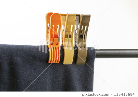 Three colorful plastic clothespins, orange, yellow, and brown, are securely holding a dark blue fabric over a horizontal metal Three colorful plastic clothespins, orange, yellow, and brown, are securely holding a dark blue fabric over a horizontal metal 135415694