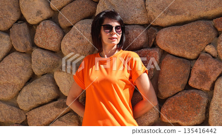 Elegant woman in orange dress standing against rustic stone wall, wearing sunglasses outdoors Elegant woman in orange dress standing against rustic stone wall, wearing sunglasses outdoors 135416442