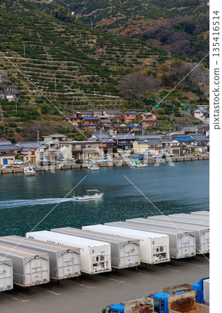 Logistics scene at Yawatahama Port in Ehime Prefecture. White container trucks and a motorboat running along a fishing boat. Port town village with Momijiyama in the background. 135416514