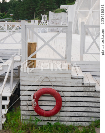 Red lifebuoy hanging on white wooden pier or beach platform with railing. Safety equipment for water rescue or emergency for summer vacation. Red lifebuoy hanging on white wooden pier or beach platform with railing. Safety equipment for water rescue or emergency for summer vacation. 135416881