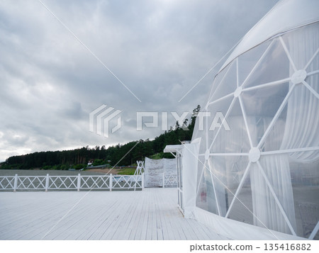 White geodesic dome on wooden deck near lake with forest landscape under cloudy sky. Outdoor glamping tent for event. White geodesic dome on wooden deck near lake with forest landscape under cloudy sky. Outdoor glamping tent for event. 135416882