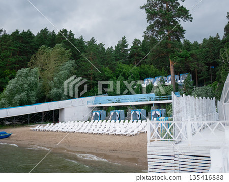 Empty beach with lounge chairs near a green forest and lake shore. Recreation area for summer vacation and travel background. Empty beach with lounge chairs near a green forest and lake shore. Recreation area for summer vacation and travel background. 135416888