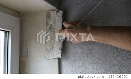 A hand with a wide trowel is leveling the surface of the slope of a small window from inside the room, smoothing the plaster on the window sill with a construction spatula. A hand with a wide trowel is leveling the surface of the slope of a small window from inside the room, smoothing the plaster on the window sill with a construction spatula. 135416961
