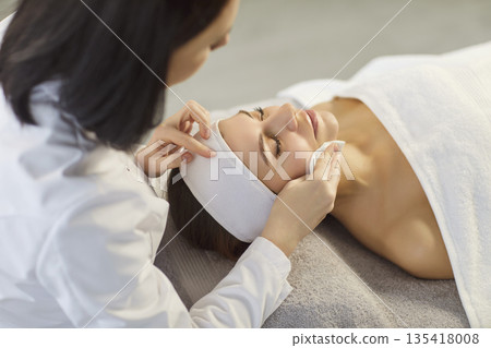 Skincare specialist cleansing woman's face before facial massage at spa 135418008
