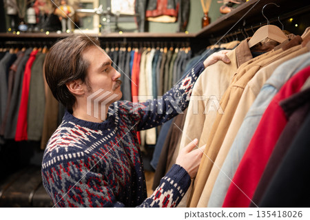 Handsome man consumer choosing clothing in vintage second hand boutique, picking jacket from hanger. 135418026