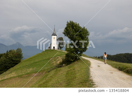 Young woman walking towards Church. The Jamnik Church is a charming 15th-century chapel in the Kamnik-Savinja Alps near Kranj, views of the surrounding mountainous landscape. The Church of St. Primoz 135418804