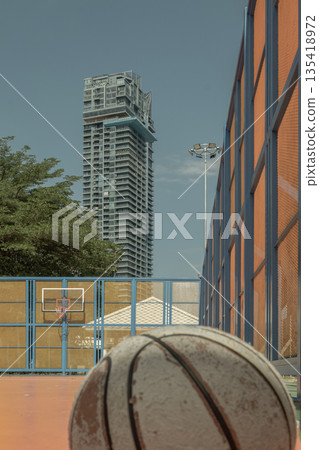Resting on the orange court surface, a worn basketball sits before a backdrop of contemporary high-rise, green trees and the sky. Resting on the orange court surface, a worn basketball sits before a backdrop of contemporary high-rise, green trees and the sky. 135418972