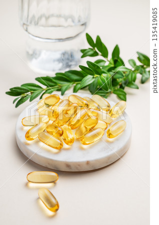 Yellow softgel vitamins capsules on a marble stand on a light background with glass of water Yellow softgel vitamins capsules on a marble stand on a light background with glass of water 135419085