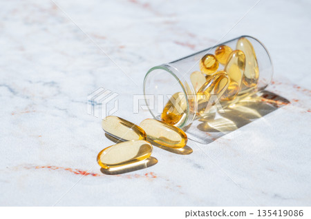 Yellow softgel vitamins capsules scattered in a bottle on a marble background close up. Yellow softgel vitamins capsules scattered in a bottle on a marble background close up. 135419086