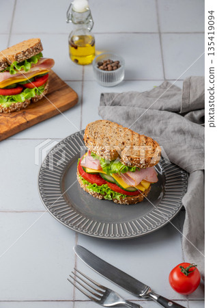 Sandwich or toast with ham, cheese, lettuce and vegetables on a plate on a gray tile background Sandwich or toast with ham, cheese, lettuce and vegetables on a plate on a gray tile background 135419094