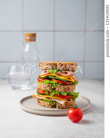 A triple sandwich with ham, cheese, lettuce, and vegetables on whole grain bread on a plate A triple sandwich with ham, cheese, lettuce, and vegetables on whole grain bread on a plate 135419095