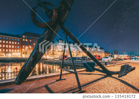 Helsinki, Finland. Old Anchor On Pier With View Of Pohjoisranta Street In Evening Night Illuminations. Night stars shining above street Helsinki, Finland. Old Anchor On Pier With View Of Pohjoisranta Street In Evening Night Illuminations. Night stars shining above street 135419172