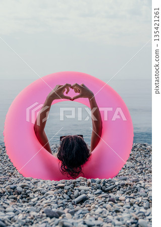 Hands heart gesture, pink inflatable ring, relaxing woman lying on summer pebble beach looking at the calm ocean. Hands heart gesture, pink inflatable ring, relaxing woman lying on summer pebble beach looking at the calm ocean. 135421261