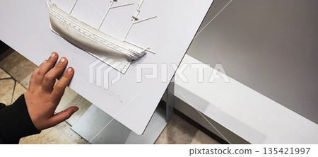 A child's hand explores Braille on a white stand depicting a three-dimensional boat. Concepts of inclusivity, sensory learning, and interactive interaction in educational or museum settings. 135421997