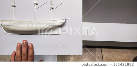 A child's hand explores Braille on a white stand depicting a three-dimensional boat. Concepts of inclusivity, sensory learning, and interactive interaction in educational or museum settings. 135421998