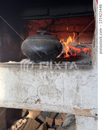 Cast iron covered with a lid. Stands on the hearth of a Russian stove. In the background, fire burns in the stove firebox. At the bottom, a woodpile with firewood is visible. Cooking food in a vintage 135423446