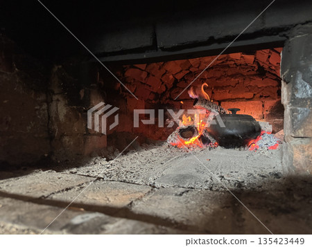 Cast iron covered with a lid. Stands in the stove firebox. Among coals and burning firewood. Cooking food in a Russian stove. Cast iron covered with a lid. Stands in the stove firebox. Among coals and burning firewood. Cooking food in a Russian stove. 135423449