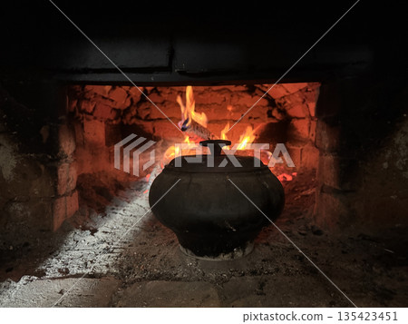 Cast iron covered with a lid. Stands on the heating stove hearth. In the background, fire burns in the stove firebox. Cooking food in a vintage stove. 135423451