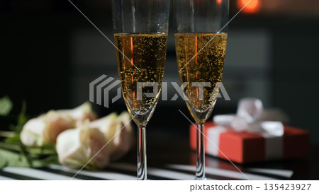 Champagne bubbles rise in glasses beside roses and a gift box at home. St. Valentines celebration concept Champagne bubbles rise in glasses beside roses and a gift box at home. St. Valentines celebration concept 135423927