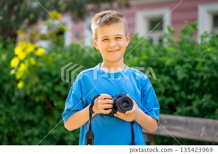 Cute boy in a blue T-shirt holds a digital camera in his hands and smiles. 135423969