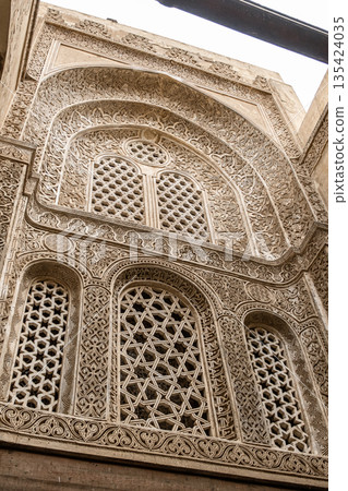 Intricate Islamic architecture of the Qalawun complex on Al-Muizz Street in Cairo. Detailed stone carvings and geometric patterns adorn the walls and windows. Intricate Islamic architecture of the Qalawun complex on Al-Muizz Street in Cairo. Detailed stone carvings and geometric patterns adorn the walls and windows. 135424035