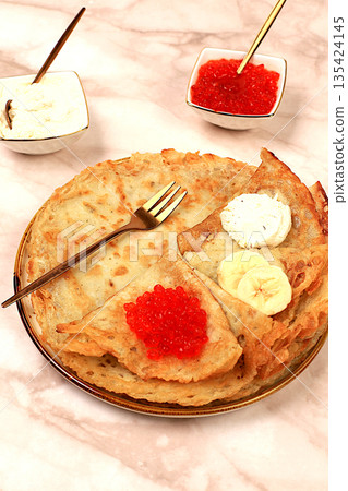 Delicious Russian pancakes with soft cheese, bananas, and red caviar on a marble table. Healthy breakfast with fresh ingredients, Maslenitsa concept and traditional Russian cuisine, selective focus Delicious Russian pancakes with soft cheese, bananas, and red caviar on a marble table. Healthy breakfast with fresh ingredients, Maslenitsa concept and traditional Russian cuisine, selective focus 135424145